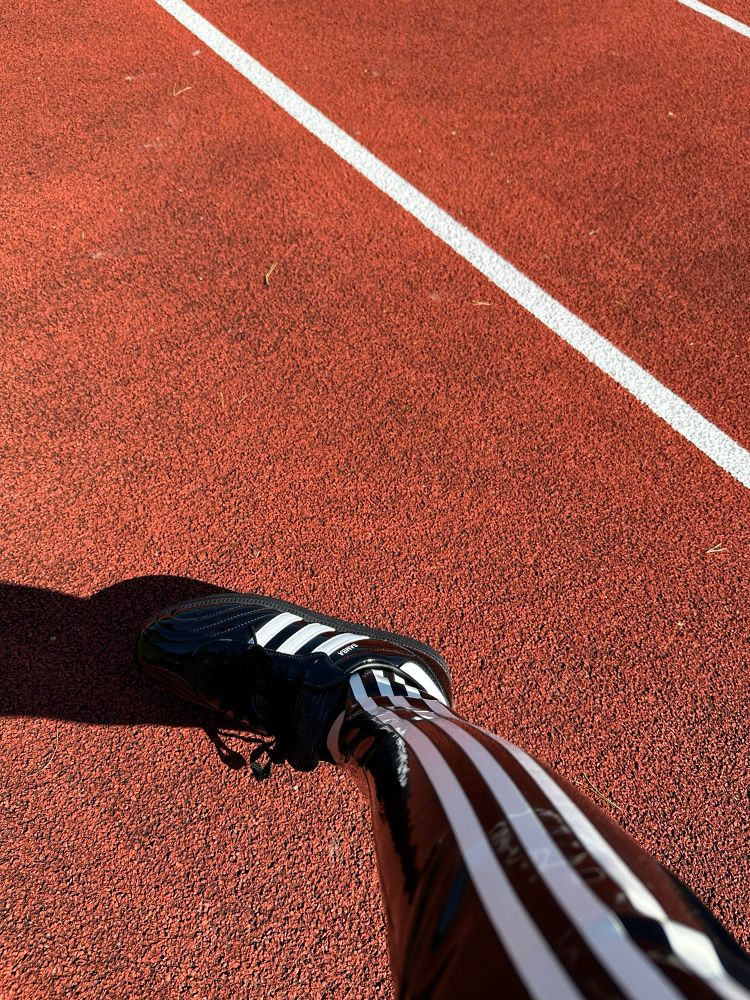 Loki in latex Adidas clothing at a running track. 

The track is red and you can see the white lines in the background. 

You can just see a very shiny black latex clad leg with white stripes down the side and a very shiny pair of Adidas running shoes too