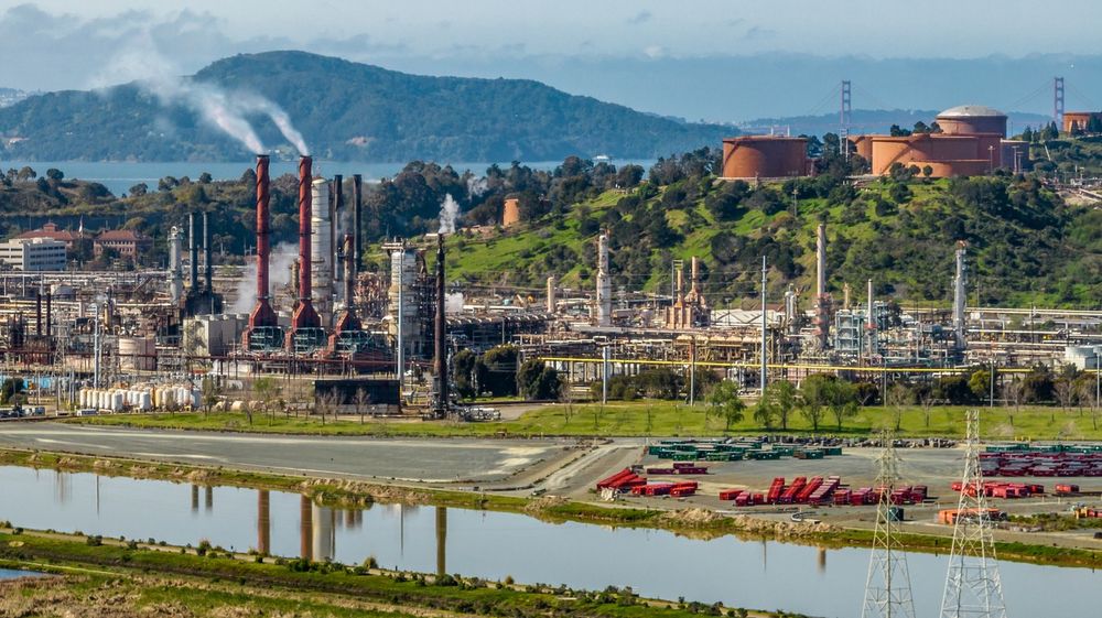 Smoke rises from smokestacks at an oil and gas refinery. The smokestacks are reflected in the water next to it. A hill and bridge can be seen in the distance.