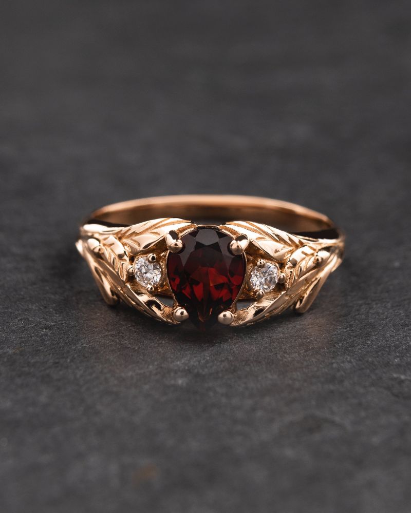 A gold engagement ring, featuring a leaf design wrapping around a pear cut garnet in the centre, and two small diamonds to either side of the gem.