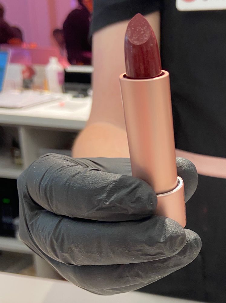 A black gloved hand holding a rose-gold tube of lipstick with a bold plummy vampy color.