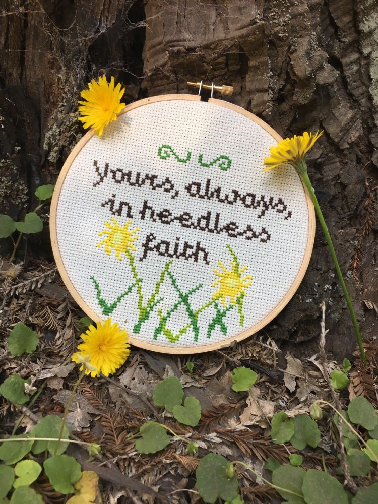 A cross stitch reading “yours always in heedless faith” with dandelion flowers