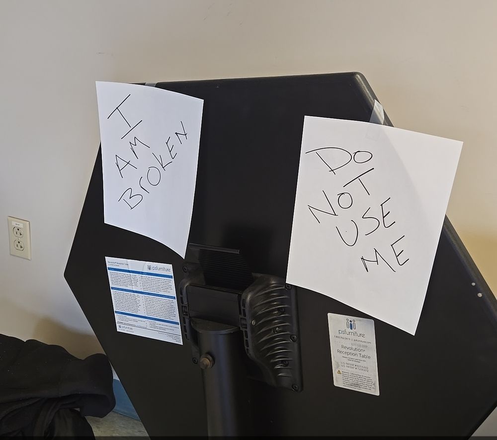 A folded hexagon shaped table with two signs on it. One says "I am broken" and one says "do not use me"