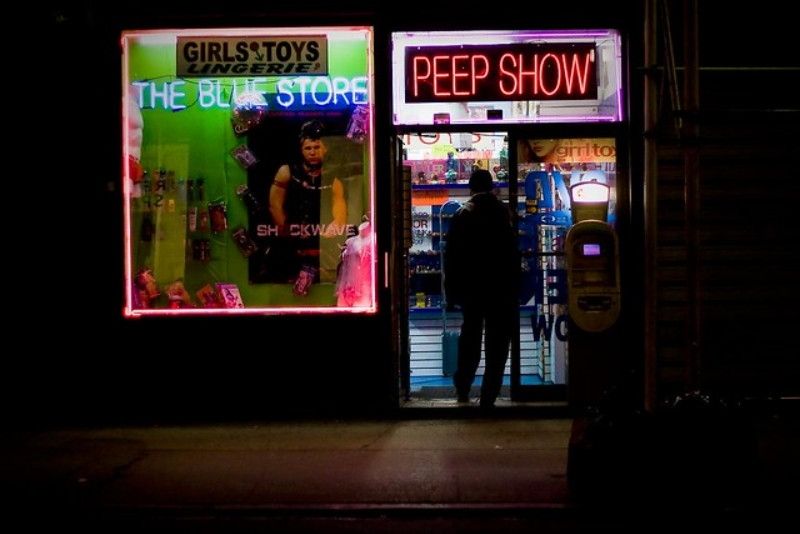 Patron walks into an adult store at night. Neon signs above the doorway and in the window. 