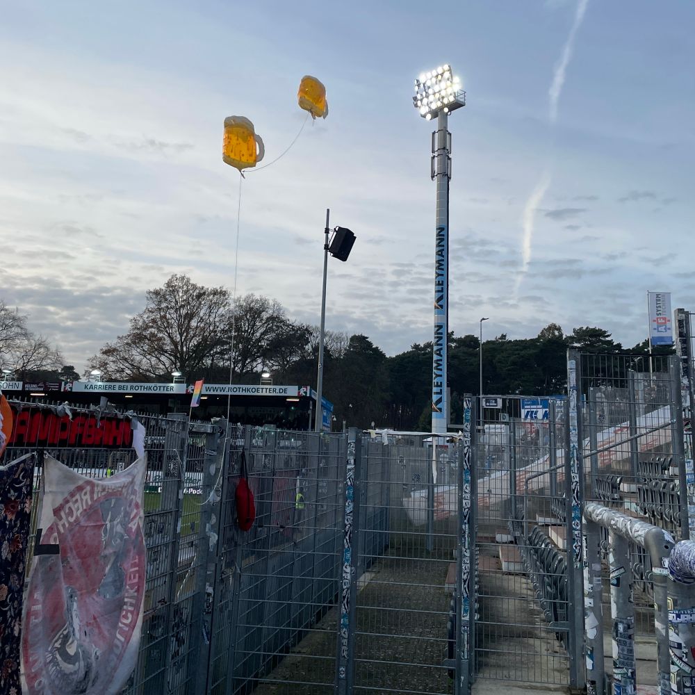 Foto aus dem Gästeblock heraus: Zaunfahnen von hinten, heliumgefüllte Bierkrugballons, Abendhimmel, Flutlicht und ein Teil der Gegengerade.