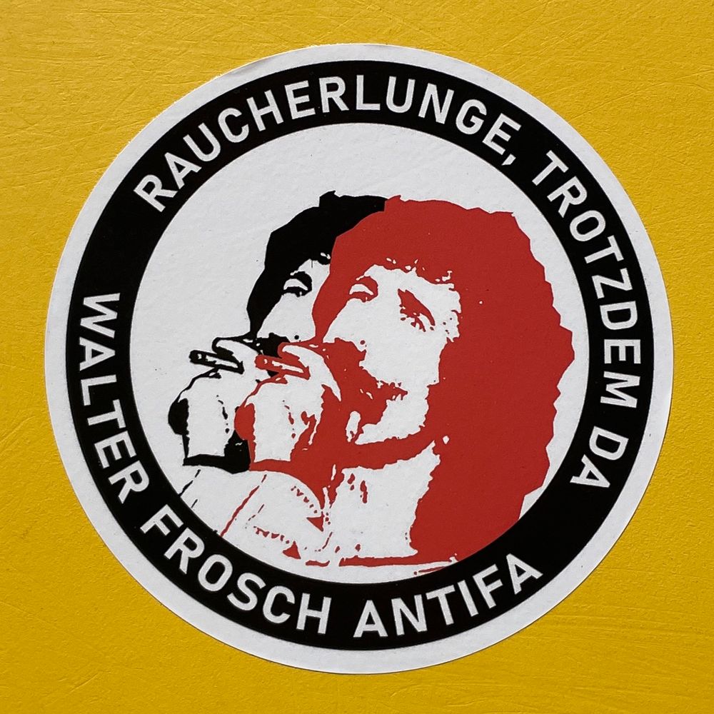 Ein Kleber auf gelbem Grund mit einem rauchendem Walter Frosch in rot und schwarz auf weißem Grund, dazu der Text „Raucherlunge, trotzdem da - Walter Frosch Antifa“.