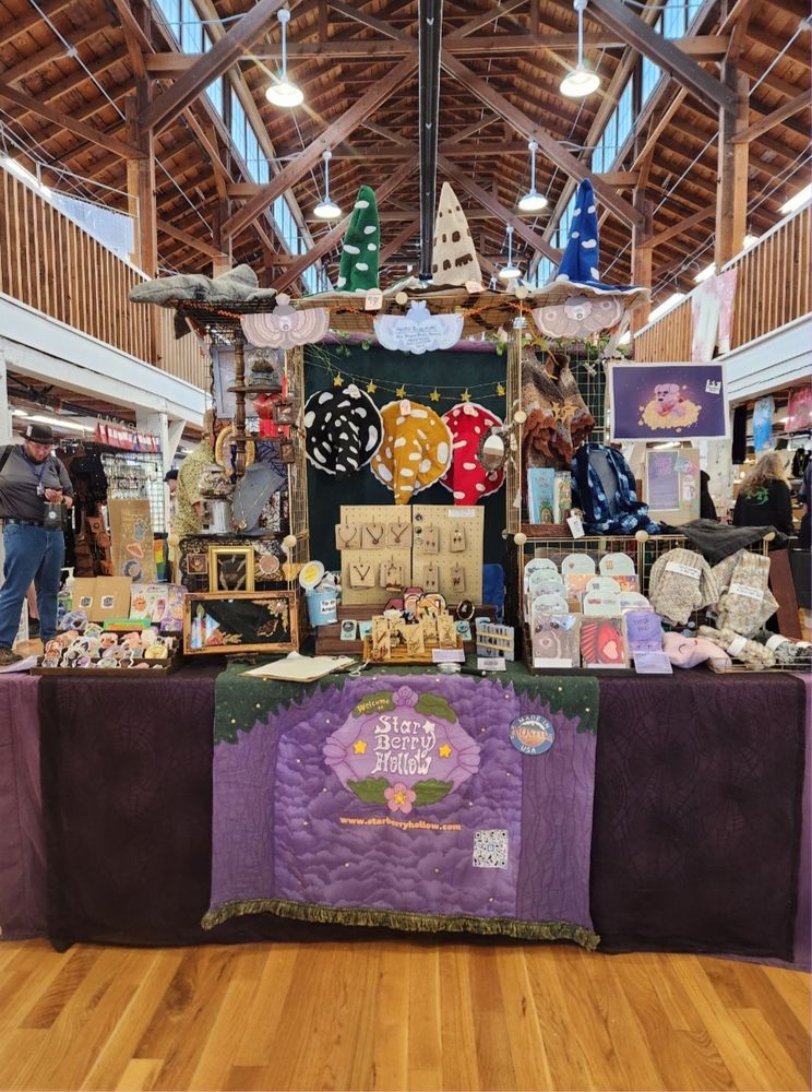 A photo of Star Berry Hollow's vendor table set up, featuring mushroom witch hats, curiosities, stickers, greeting cards, jewelery, knitwear and more.
