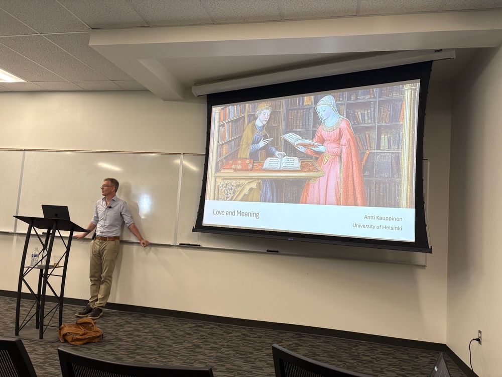Antti, a middle-aged man stands in front of a screen showing a slide with the talk’s title, “Love and Meaning”, and a medieval painting showing women holding books. 