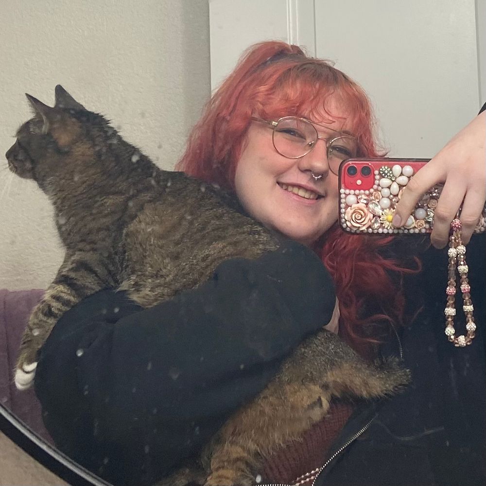 photo of my adult tabby cat named Mango and myself in front of a mirror. Feminine human with dyed dark red hair, glasses, a septum piercing, wearing a sweatshirt and holding a phone up to the mirror. i am holding Mango with one arm while she looking away from the camera. 