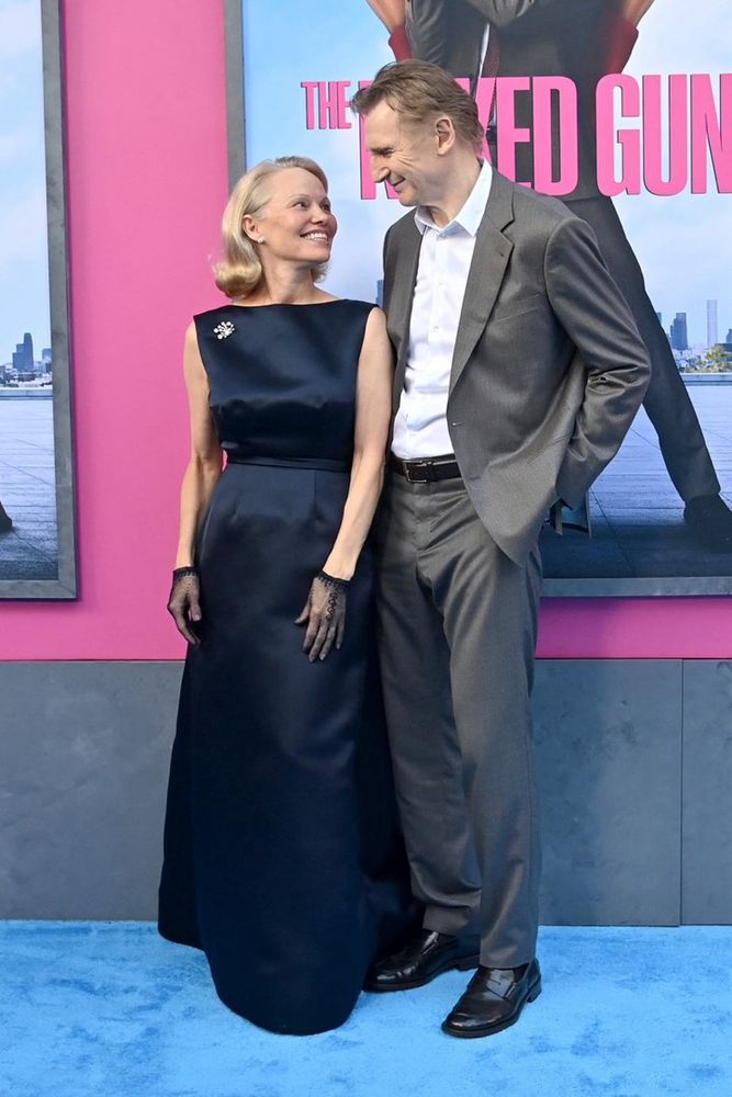 Pamela Anderson and Liam Neeson stand close together on a blue carpet, smiling warmly at each other in front of a promotional poster for The Naked Gun. Pamela wears a sleeveless, floor-length navy gown with a brooch on her left shoulder and sheer black lace gloves. Liam wears a light gray suit with an open-collared white shirt and black dress shoes. The background features bright pink elements from the movie poster, creating a bold contrast with their classic attire.
