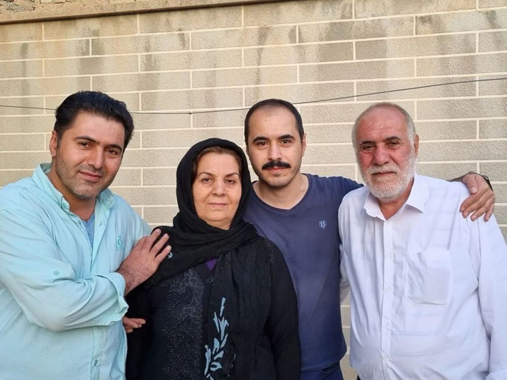 Four people stand together in front of a beige brick wall. From left to right: Hassan Ronaghi, wearing a light blue shirt, gently rests his hand on the shoulder of his mother, who is dressed in a black headscarf and dark clothing; next to her is Hossein Ronaghi, with a shaved head and mustache, wearing a navy blue T-shirt; on the far right is their father, an older man with a white beard and short white hair, dressed in a white shirt. The family stands close together, appearing somber but united.