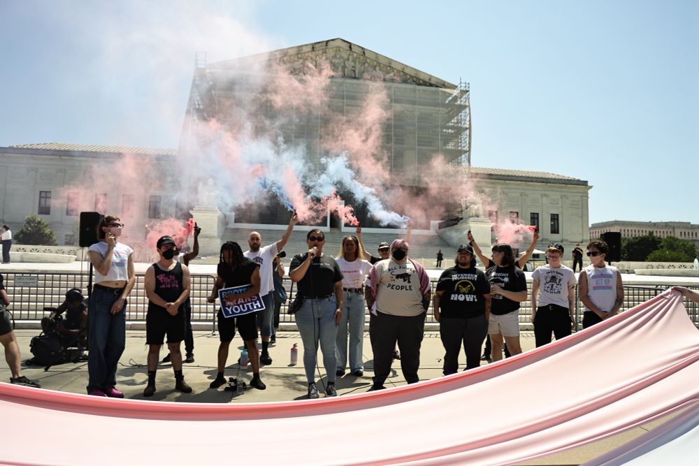 Activists release blue pink and white smoke in front of the SCOTUS with the trans flag colors draped in front of them