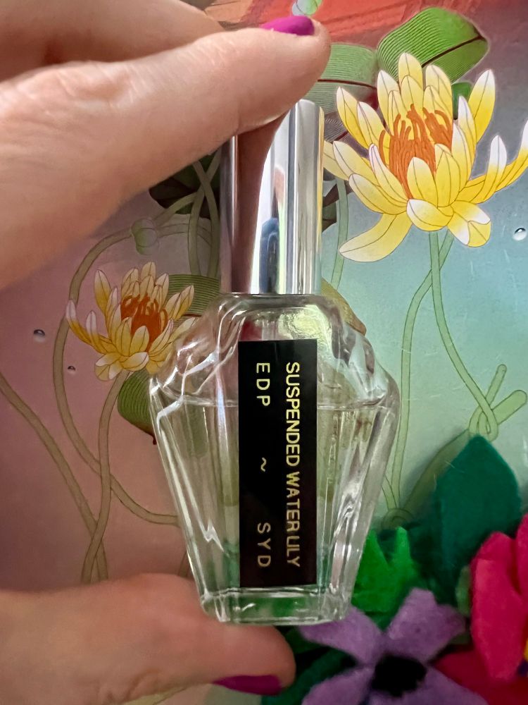 A photo of a small bottle of the perfume Suspended Water Lily by SYD Botanica, held up against a backdrop of art from the brand. 