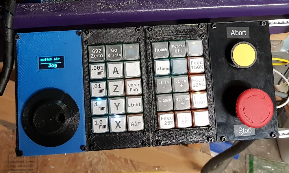 A shop-built CNC control panel, with a dial and display on the far left, to the right is two panels of 3x5 buttons with custom labels.  You can see letters signifying axis on some, and length increment on others.  

Further right from this is an abort button and am estop switch.