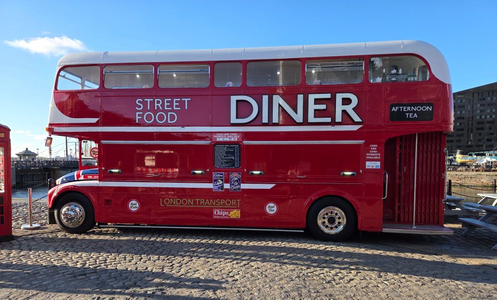 Red double decker bus that doubles as a diner. On the side are the words Street Food Diner in white, as well as London Transport in gold. Afternoon Tea is written in white on a black sign.