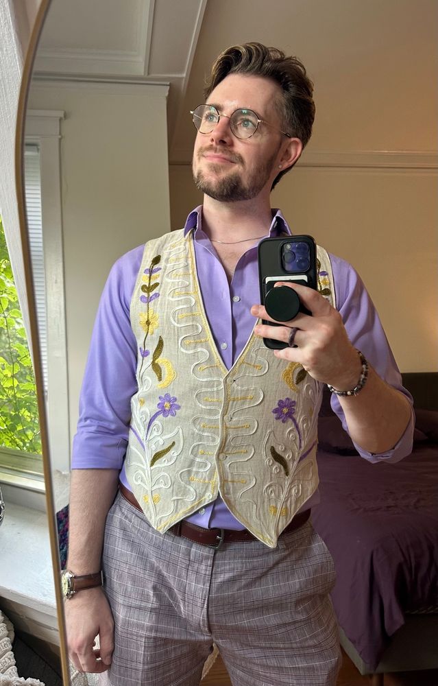 mirror selfie showing me wearing glasses looking out of frame behind the mirror, wearing a creme floral vest over a lavender shirt and purple plaid shorts