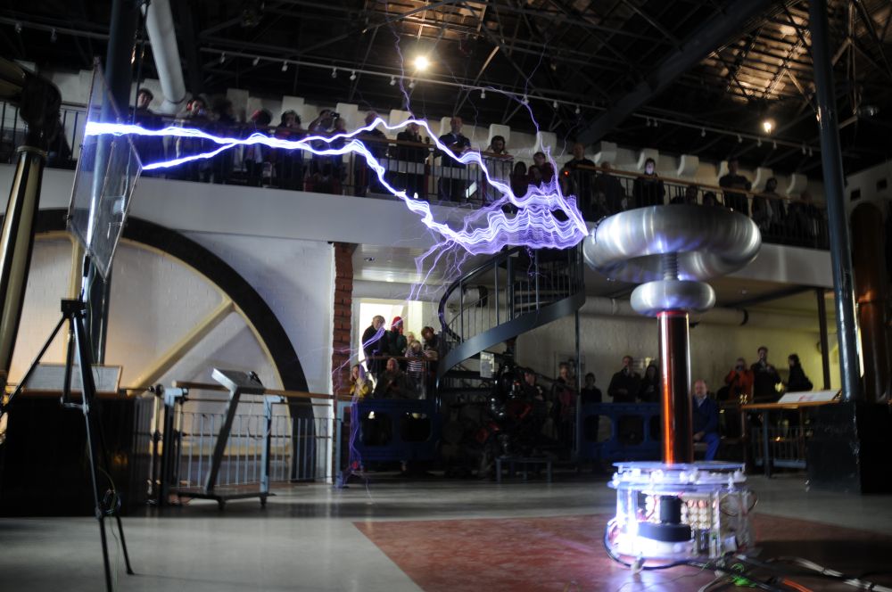 Tesla coil striking a mesh target. Onlookers from a balcony and around the room, behind barriers. 