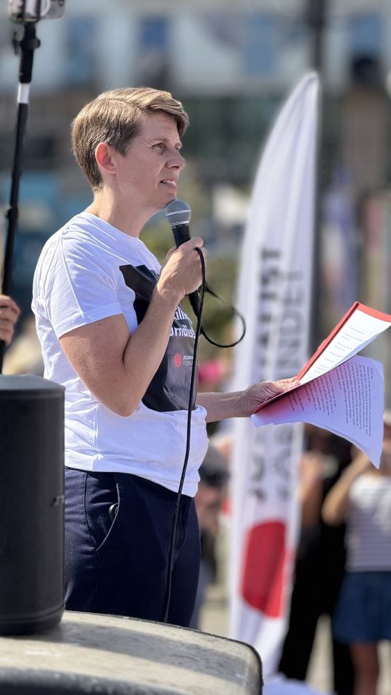 Auto-generated description: A person is speaking into a microphone while holding a red folder, with a banner visible in the background.