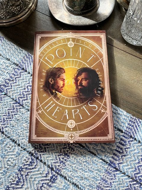 A copy of the novel Point of Hearts, with a cover showing the heads of a brown-haired man and a dark-haired man facing each other on a gold background marked with a white oval that resembles the circle of a horoscope. The novel is lying on a wooden table with a blue and white cloth and a glimpse of silver dishes in the background.
