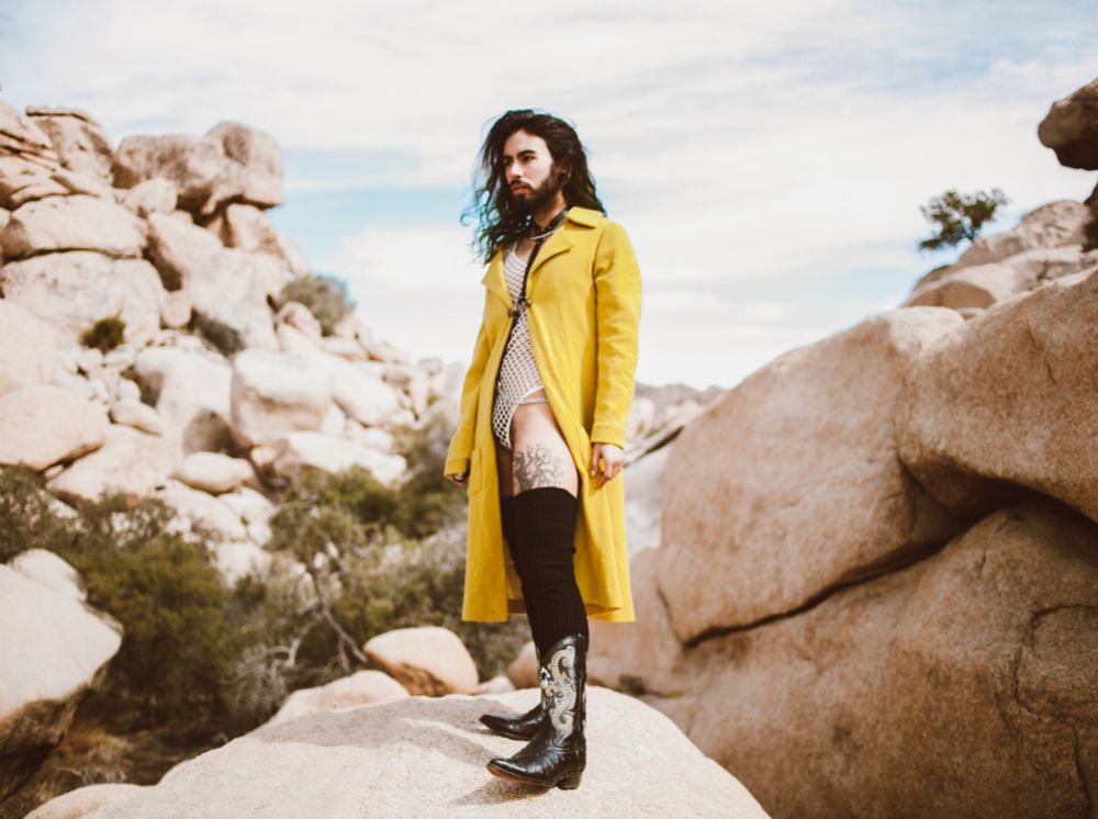 Cosmo is standing on a rock, surrounded by granite rock formations and desert plants blurred in the background. They are looking off to the side, hair gently flowing in the wind. They are wearing a white mesh bodysuit with a long yellow jacket pulled to the side, and black cowboy boots with thigh high black socks. Their stance is powerful yet somehow they also look soft.