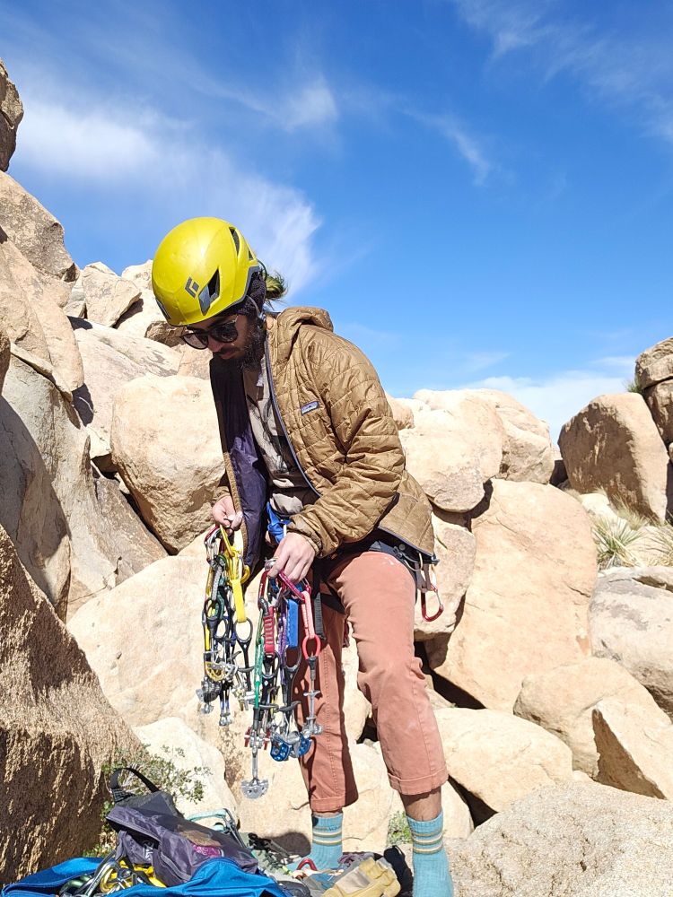 A person wearing a yellow helmet, brown puffy jacket, and sunglasses is seen holding a handful of climbing cams. It looks like they are trying to decide what to take. The background is a bright and sunny desert rockscape.