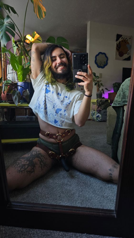 Cosmo is seen kneeling in front of their mirror taking a mirror selfie. Their legs spread, wearing a beautiful brown leather strap on, and weilding a firm black dildo. They have an elbow up with their arm gently curled behind their head, their cropped t-shirt exposing their belly. They are smiling cutely.