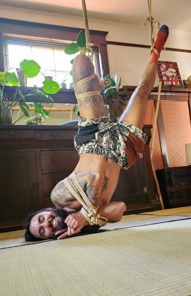 Cosmo, a bearded nonbinary person, is seen with only their head and one shoulder laying on the floor while they are being suspended in rope by a futomomo on one leg and an ankle cuff on their other leg. Their arms are bound with their hands to their chest and they are holding up a peace sign and smiling. 