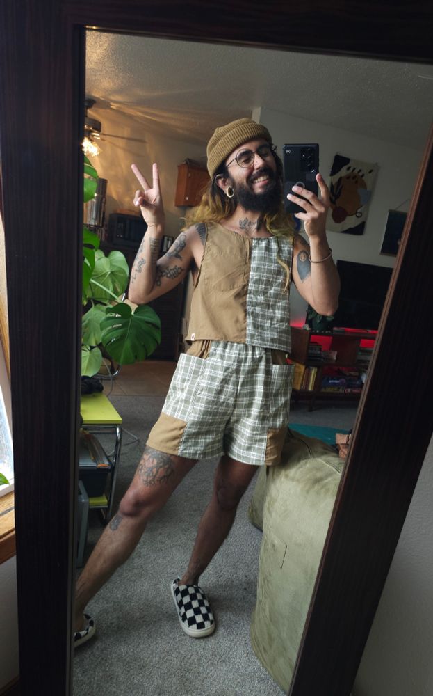 Cosmo is seen taking a mirror selfie in an outfit they sewed themselves. They are wearing a tank top that is colour blocked straight down the center, with one side being a light brown and the other side being a white and green flannel. Their shorts are the same fabrics but colour blocked with the brown being on the side panels.

Cosmo is smiling and holding up a peace sign. They have a fluffy beard, glasses, and a matching brown beanie. One of their mini braids gently dangles across their chest.