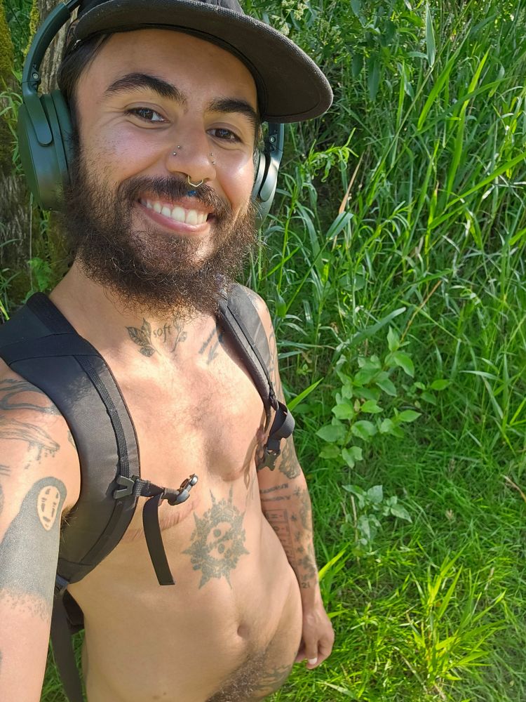 Cosmo, a bearded and tattooed individual, is seen standing nude in front of some lush grass. They have a black backpack on with the sternum strap unclipped. Cosmo is only wearing a black panelled hat and forest green over the ear headphones. They are smiling, and the sun is kissing their skin. They are glistening.