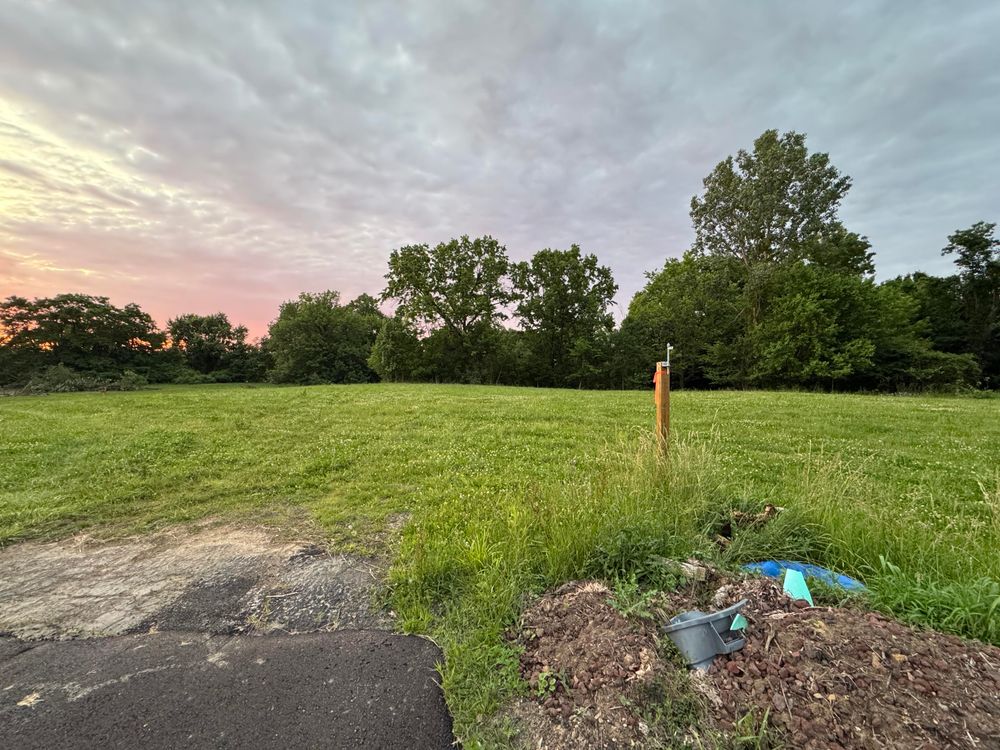 A large green grassy area, with an asphalt driveway that abruptly ends. A post and some debris in the foreground. 