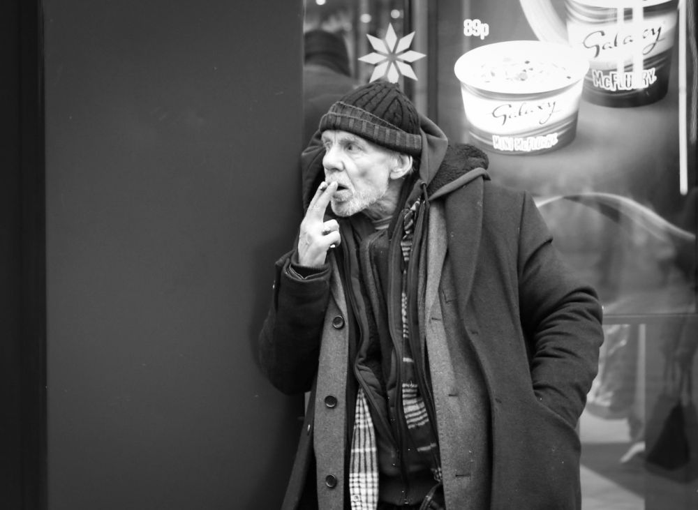Black and white image shows a well-wrapped man pulling on a hand rolled cigarette, the hand he's using to snoke holds a lighter in it. He's looking deep in thought as he stares off to his right. A star on the cafe door behind him looks like a pom-pom for his hat.
He has 4 or possibly 5 jackets on but all open at the front signalling it's warmish now but likely to get much colder later on. He is an older man, a week's worth of beard growth and a wooly hat on
