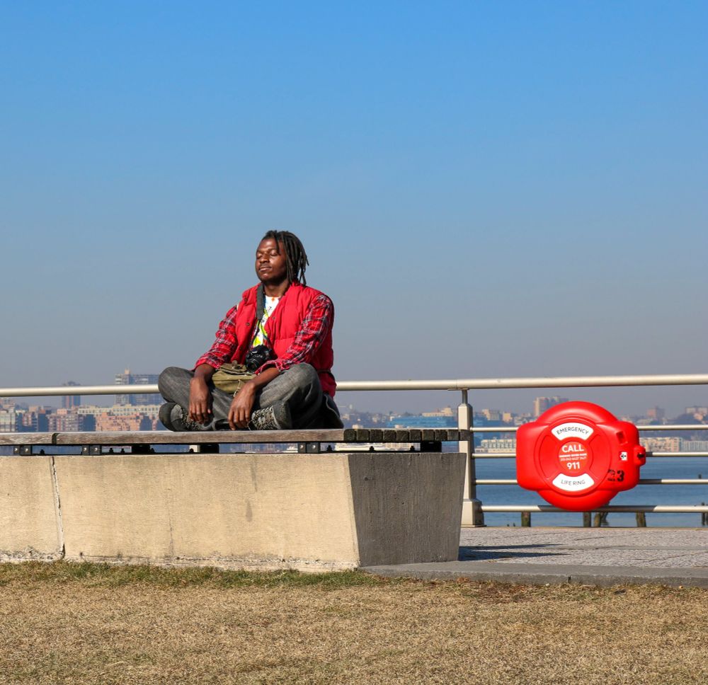 Colour image shows a photographer sat cross legged on a raised concrete bench with wooden top that starts center image and leads horizontally to the left leaving open space at the end of the bench meaning you can see the Hudson River behind them and a life ring on the metal barrier between. The photographer is wearing dark, faded gray jeans, dark sneakers and a red check jersey - they have a Canon camera hanging from their neck in front of them.