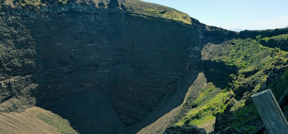 The image depicts a close-up, detailed view of the Vesuvius crater, showcasing its steep, dark, and jagged interior walls, along with some green vegetation on the outer slopes.
Crater Interior:
The dominant feature is the vast, deep depression of the crater, characterised by its dark, almost black, rocky walls. These walls reveal visible layers and textures, indicating the volcanic rock formations.
Slopes and Debris:
The bottom of the crater appears to be filled with loose rock and debris, forming a sloped, uneven floor.
Vegetation:
Patches of vibrant green vegetation are visible on the upper, outer rim of the crater and extending down some of the less steep slopes, contrasting with the dark volcanic rock.
Perspective:
The viewpoint is from the rim, looking down into the crater, providing a sense of its immense scale and depth. A wooden post is partially visible in the lower right corner, indicating a viewing area walkway for visitors.
Lighting:
The scene is well-lit by natural daylight, highlighting the contours and textures of the crater walls and the surrounding landscape