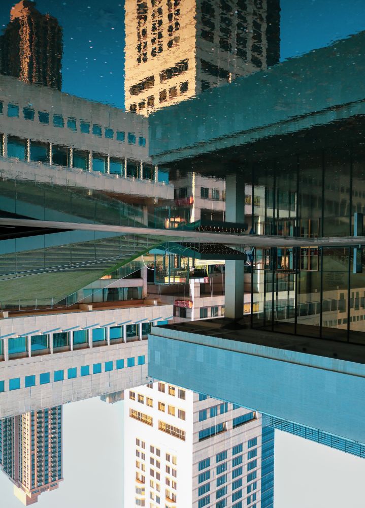 The image depicts a reflection of modern city buildings in a body of water, creating a striking symmetrical effect.
Subject Matter:
The primary subjects are tall, contemporary buildings with numerous windows, reflecting the sky and surrounding structures.
Composition:
The image is vertically oriented, with the real buildings in the lower half and their mirrored reflections in the upper half, divided by a clear horizontal line representing the water's surface.
Colour Palette:
The dominant colours are cool tones of blue and grey from the sky and building facades, contrasted with warmer tones of brown and gold from some building materials and the reflected sunlight.
Visual Effect:
The reflection is remarkably clear, almost perfectly mirroring the architectural details, creating an illusion of two identical cityscapes meeting at the waterline. This creates a sense of depth and an interesting play between reality and illusion.