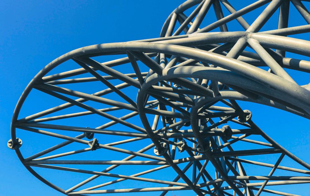 The image captures a close-up, upward-angled view of a large-scale metal sculpture, likely a public art installation, set against a clear, vibrant blue sky. The sculpture is composed of numerous interconnected grey metal beams and tubes, forming a complex, open, and curvilinear lattice structure that creates a sense of depth and intricate design. The strong sunlight casts distinct shadows within the sculpture's framework, highlighting its three-dimensional form and the interplay of light and shadow.