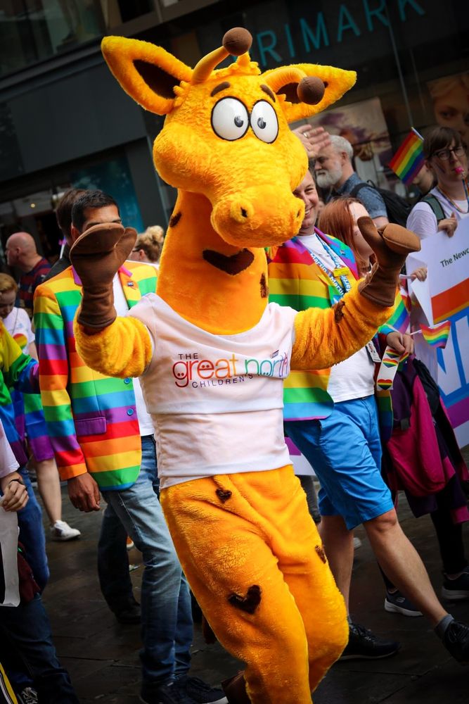 The image shows someone in a bright yellow giraffe costume with hearts instead of spots on its coat. They're wearing a white tee shirt with great north children's hospital on the front. They're walking from left of shot to right, among a crowd of other revellers in rainbow coloured outfits blurred into the background 