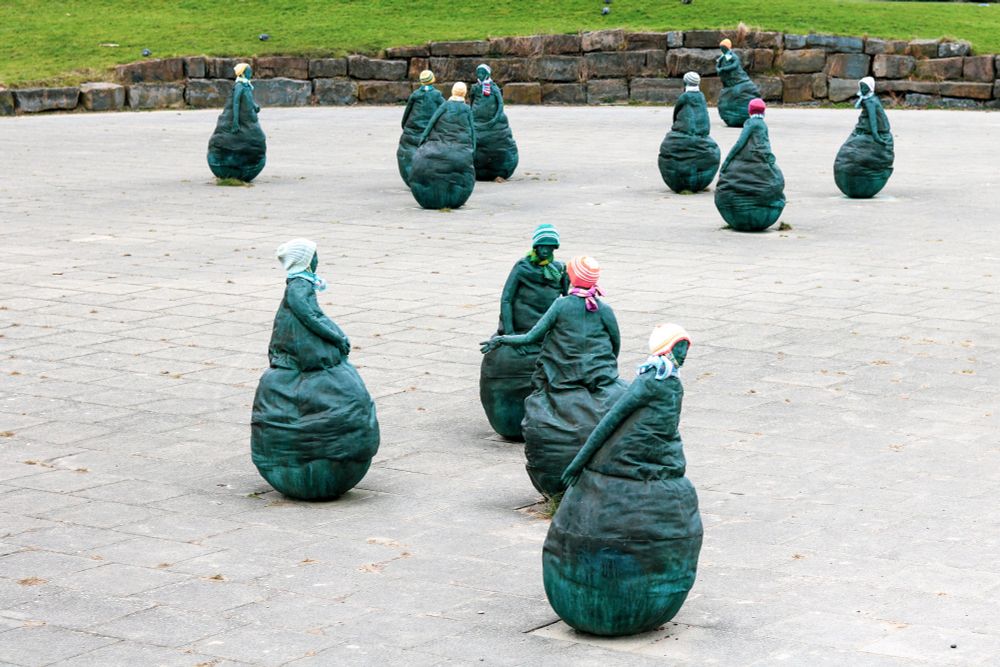 The image shows part of a public art installation known as "Conversation Piece" by Spanish artist Juan Muñoz, located near Littlehaven Beach in South Shields, UK. 
The installation comprises 22 bronze figures, each approximately 1.5 meters high and weighing about a quarter of a ton. 
Locals affectionately refer to the figures as the "Weebles" due to their rounded bases, reminiscent of the 1970s toy. 
The figures are arranged in small groups, some appearing to engage in conversation, while others gaze out to sea, reflecting the piece's title. 
In a recent development, hats and scarves were added to the figures during the winter of 2024-2025 by a local craft group member, Sheila Young, who felt the figures looked cold and wanted to honor men lost in war. 