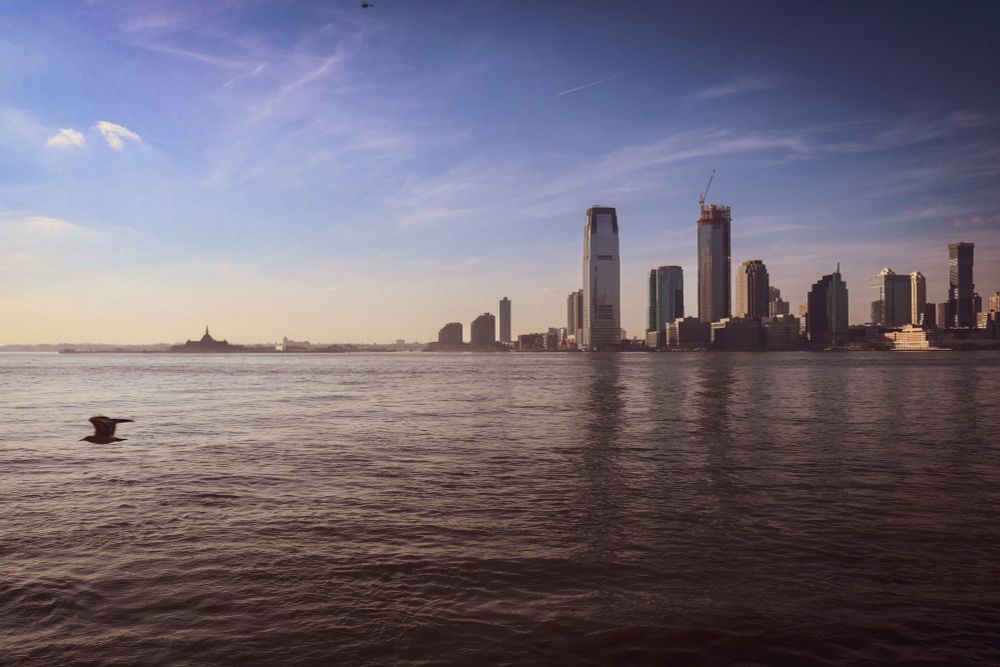 The image captures a panoramic view of a New Jersey skyline across a body of water, the Hudson River, with a prominent foreground of dark, rippling water. The skyline features a cluster of modern skyscrapers, including one particularly tall building and others in various stages of construction, suggesting urban development. On the far left, a distinct, lower silhouette of Ellis Island Museum with a structure resembling a fort or historical building is visible in the distance. The sky above is a mix of clear blue and scattered clouds, with hints of warm light, suggesting near sunset. A solitary bird is captured in flight over the water in the lower left portion of the image.