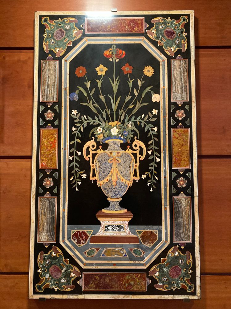 Photo of a stone painting featuring a bouquet in a vase in the center, surrounded by a border with a geometric pattern. 