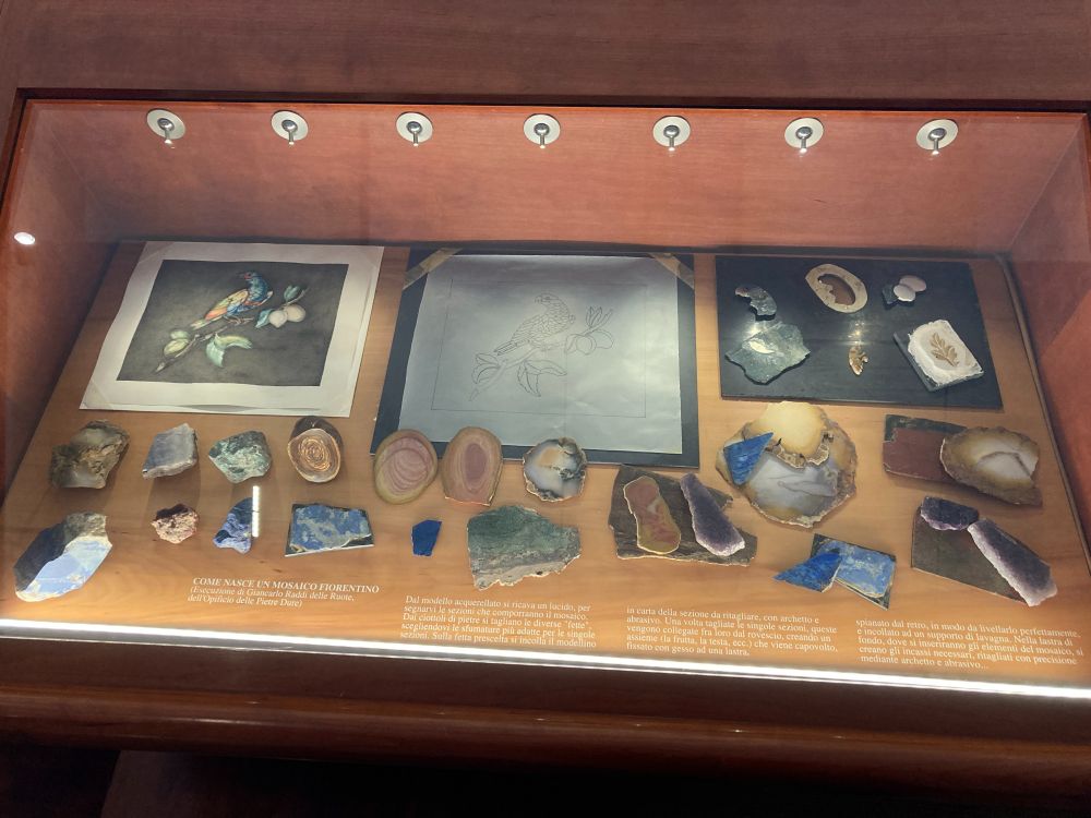 Photo of a display case that shows the different stages of making a pietra dure, including the initial watercolor, model drawing for transfer, small sections of stones that are joined to create part of the image, and different kinds of stones as the color palette. 