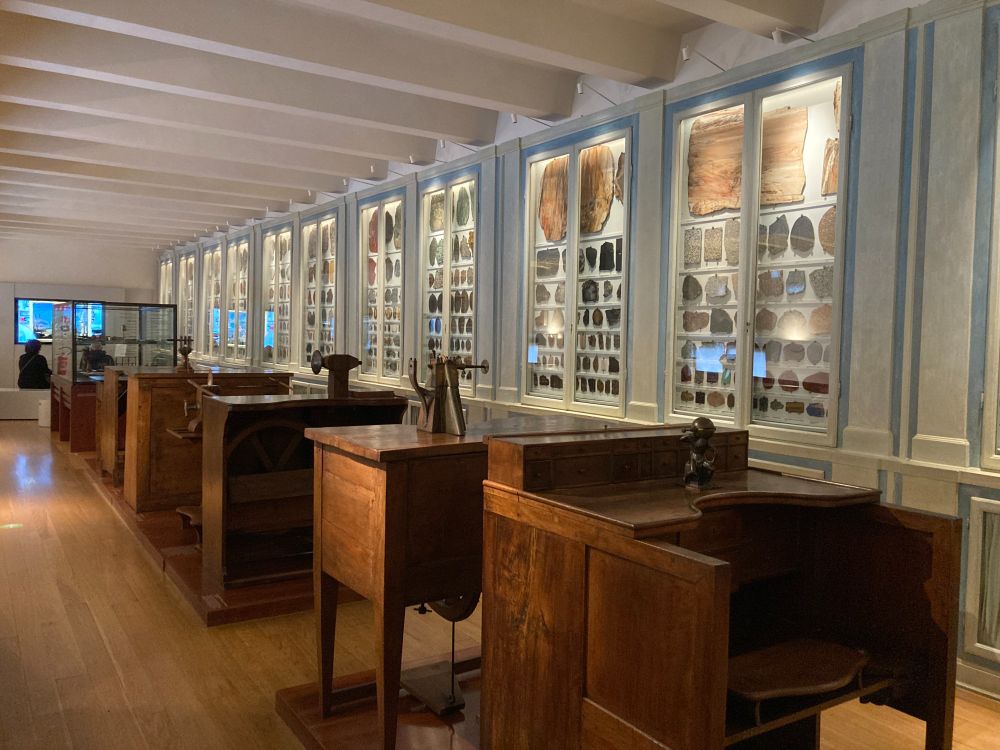 Photo of a section of the museum, showing historical workstations for cutting, grinding, and polishing stones, with a wall-to-wall display of different kinds of precious stones used for pietre dure in the background.