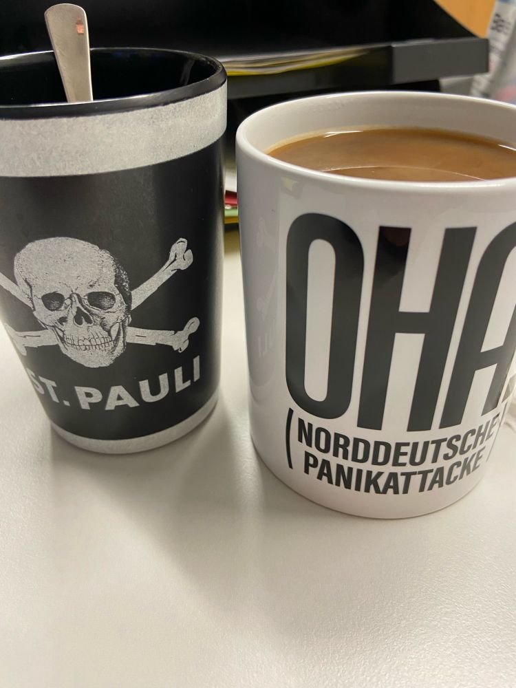 Schwarzer Becher mit Aufdruck Totenkopf und Schriftzug St. Pauli steht auf einer weißfarbigen Tischplatte. Daneben steht ein weißer Keramikbecher mit der Aufschrift „Oha“. Darunter in Klammern „Norddeutsche Panikattacke“. Der Becher ist mit Kaffee gefüllt.