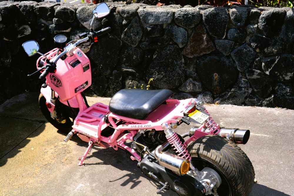 A bright pink motorcycle, the licence plate suggests is a moped but has an unusual design, parked against a stone wall. 