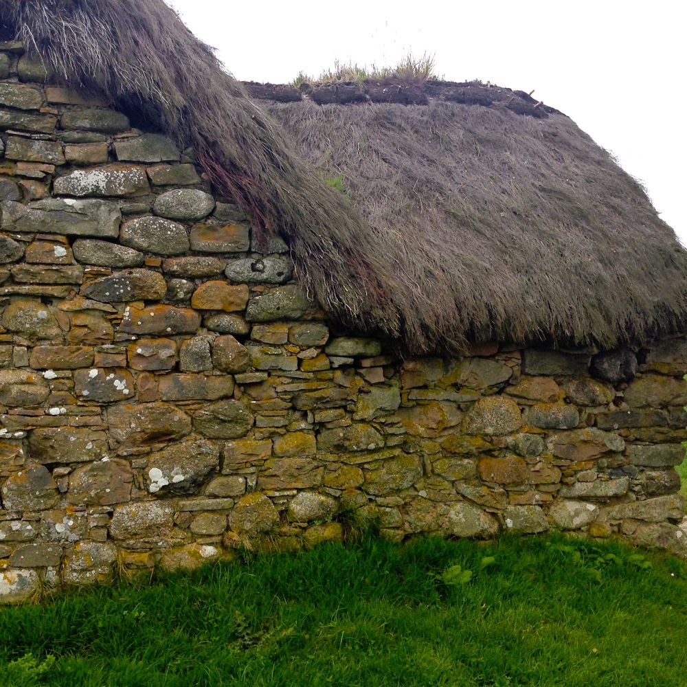 A rear view of part of a tiny ancient cottage comprised of many different shaped rocks. The roof is thatched and higher, sloping down onto the right as the picture is viewed. The grass at the bottom is lush and vivid green.