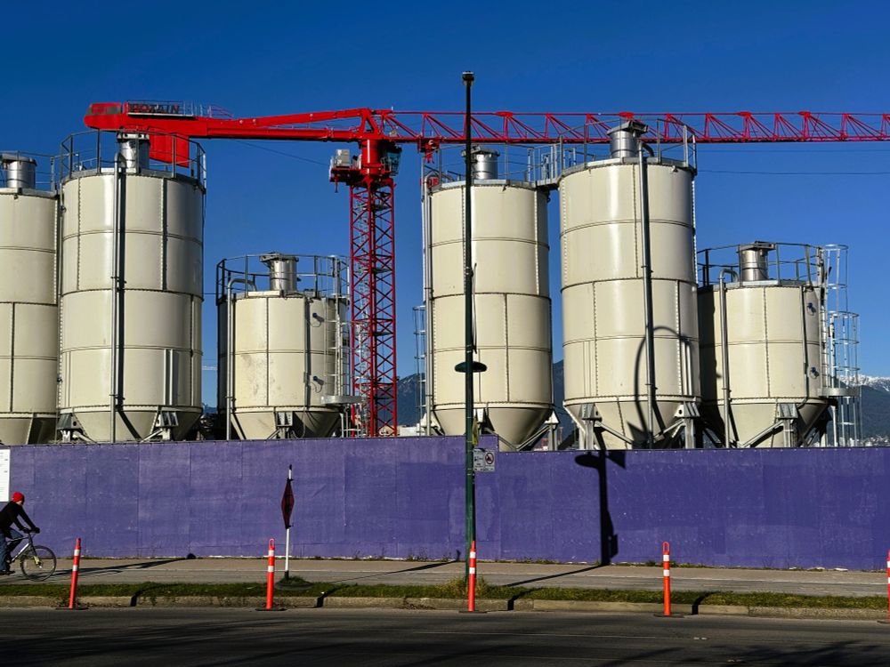 A construction site with six off white silos used, I guess, for holding chemicals above which is a fire engine red crane against a deep blue sky. There is a purple mesh fence enclosing it off the street, a single extremely tall lamp post just off centre, a cyclist with a red hat has partially entered from the left on the sidewalk with four red and white bollards and a black flag on a metal pole. There is a glmpse of snow coloured mountains in between the silos.
