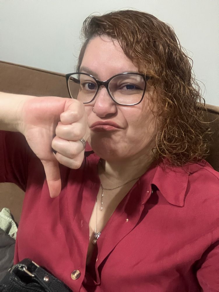 Selfie colorida interna. 
Sou uma mulher de pele bege clara, cabelo cacheado ruivo claro dourado, logo abaixo do queixo, uso óculos de armação escura, retangular grande. Estou fazendo uma careta e tenho a mão à frente do rosto, fazendo sinal de negativo com o dedão pra baixo. Uso um vestido-camisa cor de vinho.