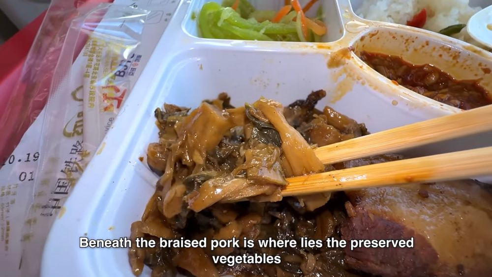 a screenshot of a youtube video where someone is picking up vegetables cooked in a sauce with chopsticks in a bento box meal served on a train. the caption reads “Beneath the braised pork is where lies the preserved vegetables”
