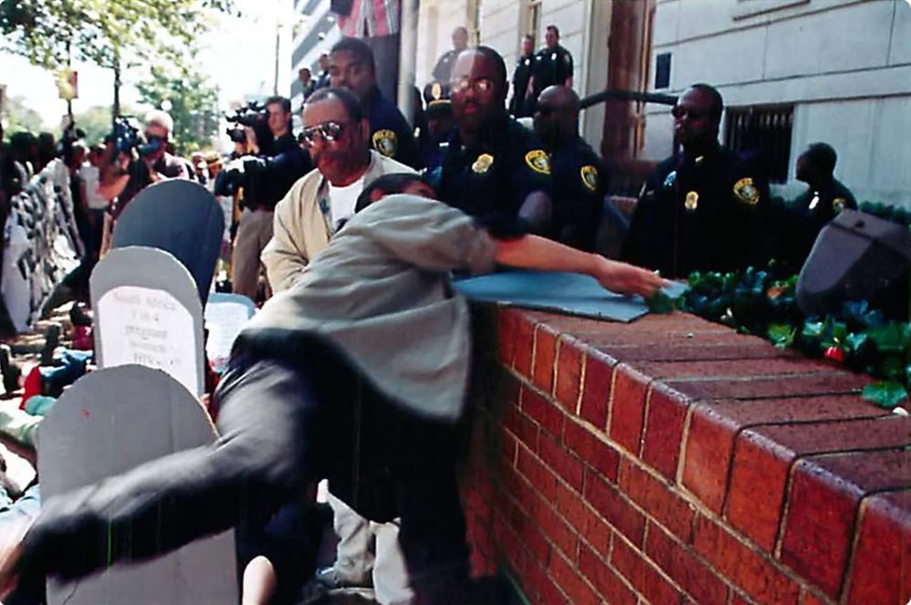 "Queer illder ancestor Kiyoshi Kuromiya of ACT UP Philadelphia starts to hurl himself
onto brick wall outside the U.S. Trade Representative’s Office
in 1999 at his final civil disobedience action
after a life of justice struggles,
at a protest for global HIV treatment access."