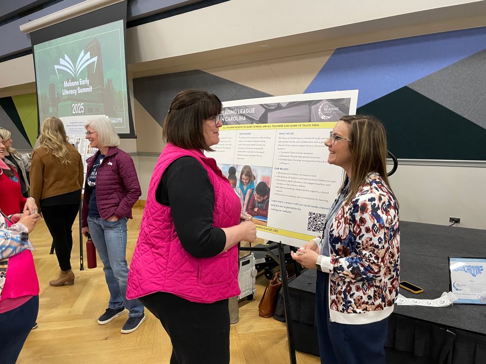 Linda stands in front of the TRL-NC poster speaking to an audience member. 