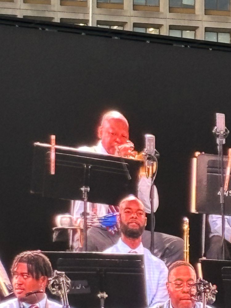 Photo of Wynton Marsalis playing solo trumpet surrounded by fellow musicians. Stark black background. All musicians visible in shot are in matching white shorts, striped ties and light brown pants.