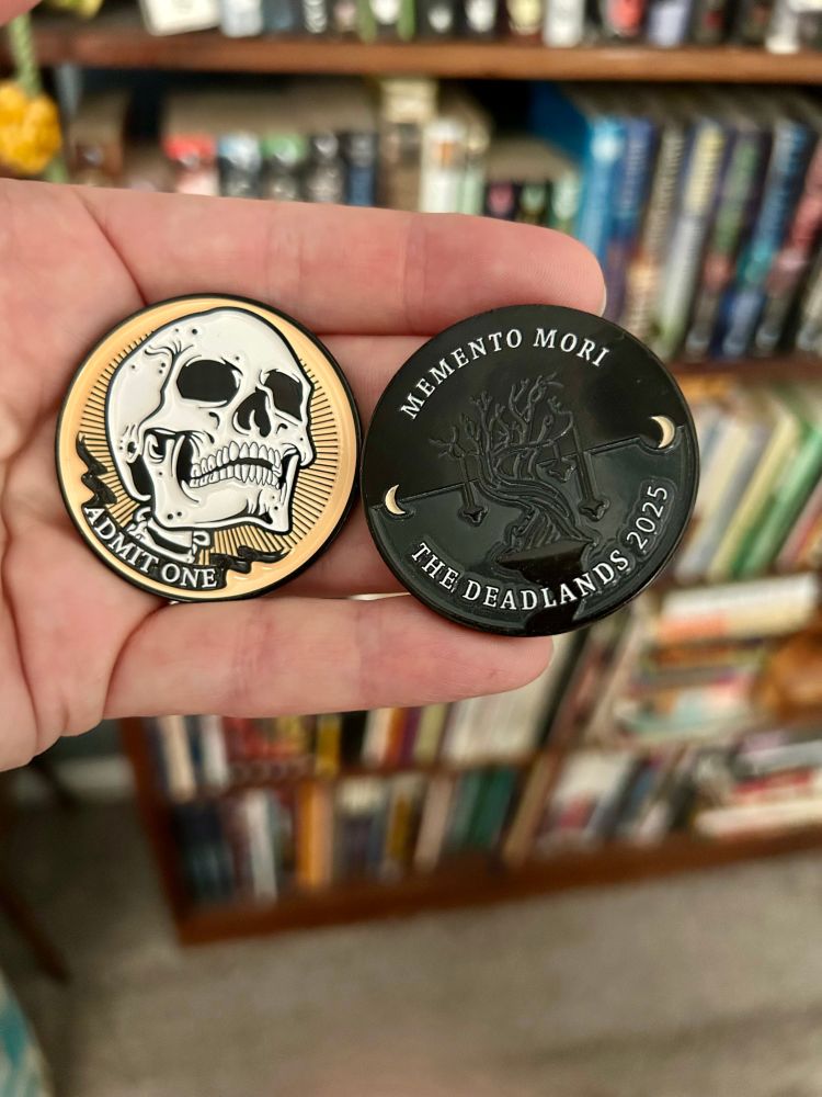 Two previous coins from The Deadlands. The left one is a skull on a bright mustard yellow background, looking up and to the left. A banner below the skull reads "ADMIT ONE". The right one has a dark tree on a black background. The text on it reads "MEMENTO MORI" and "THE DEADLANDS 2025".